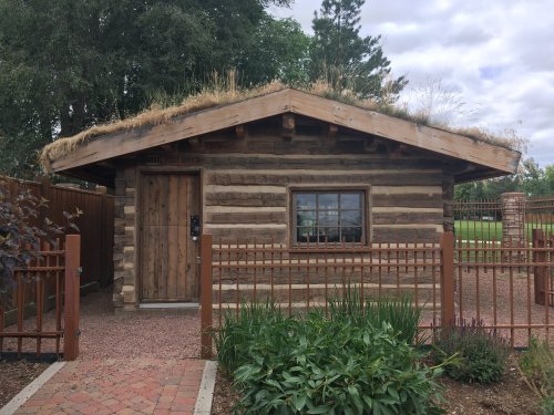 A small log cabin with a grass-covered roof, surrounded by greenery and a wooden fence.