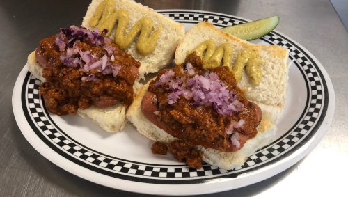 Two hot dogs topped with chili, diced onions, and mustard, served on white bread with a pickle on the side.
