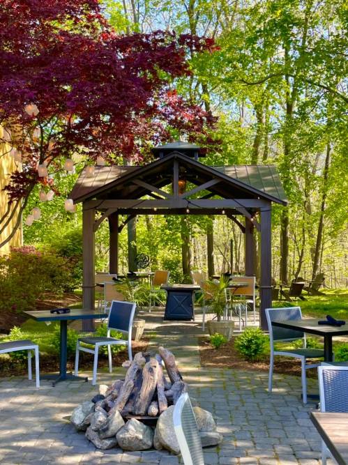 A cozy outdoor gazebo surrounded by trees, with tables and chairs arranged around a stone fire pit.
