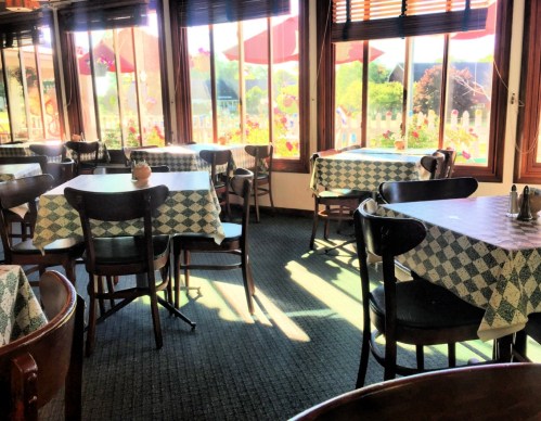 Bright restaurant interior with empty tables, checkered tablecloths, and large windows showcasing a sunny outdoor view.