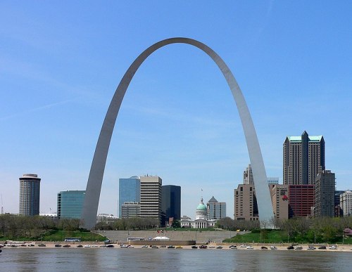 The Gateway Arch towers over the St. Louis skyline, with the Mississippi River in the foreground and buildings behind.