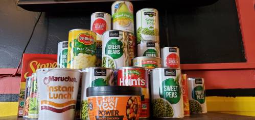 A pyramid of various canned foods, including vegetables, soups, and instant noodles, on a colorful shelf.