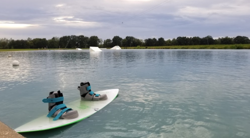 A wakeboard with boots sits on the edge of a calm, blue water surface, surrounded by greenery and a cloudy sky.