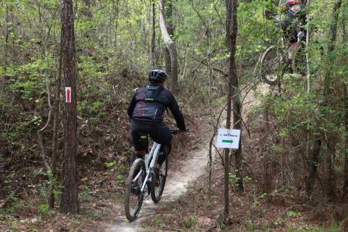 A mountain biker rides along a narrow trail in a wooded area, with a sign indicating the path ahead.