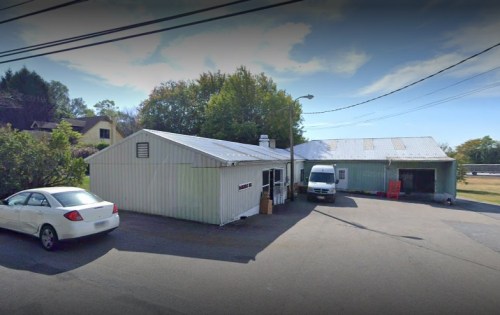 A small, light-colored building with a metal roof, surrounded by trees and parked vehicles on a sunny day.