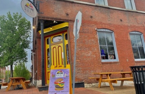A colorful restaurant entrance with a yellow door, brick facade, and outdoor seating area. A sign advertises food.
