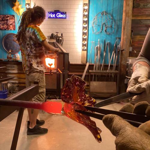 A glassblower shapes molten glass in a workshop, with vibrant colors and tools visible in the background.