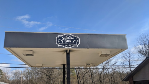 Sign for "Smoking Gun" on a gas station canopy, with a clear blue sky and trees in the background.