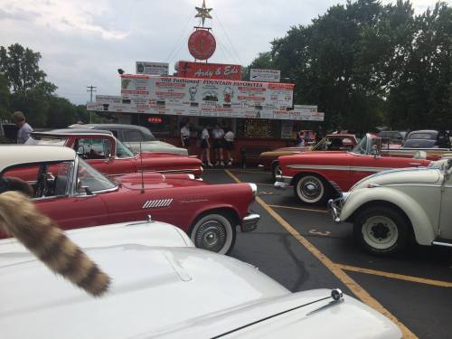 A vintage car gathering in front of a retro diner, with classic cars parked and people ordering food.
