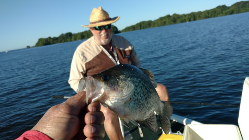A person holds a fish in the foreground, while another person smiles in the background on a sunny lake.