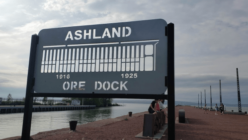Sign for Ashland Ore Dock, with dates 1916 and 1925, overlooking a calm waterway and a pier in the background.