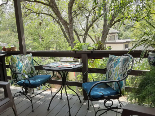 A cozy outdoor balcony with two chairs and a small table, surrounded by lush greenery and trees.