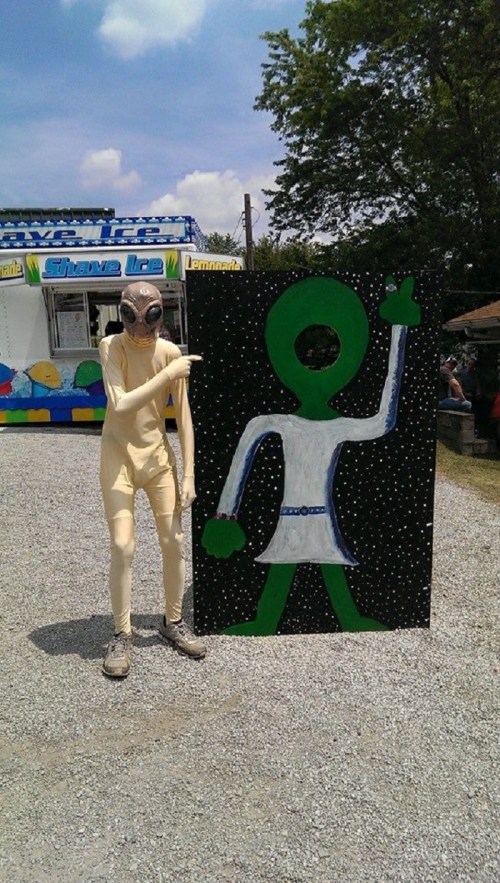 A person in an alien costume poses next to a painted alien figure on a black backdrop with stars.