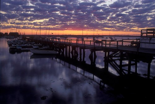 A serene sunset over a calm harbor, with boats docked and a wooden pier reflecting in the water.