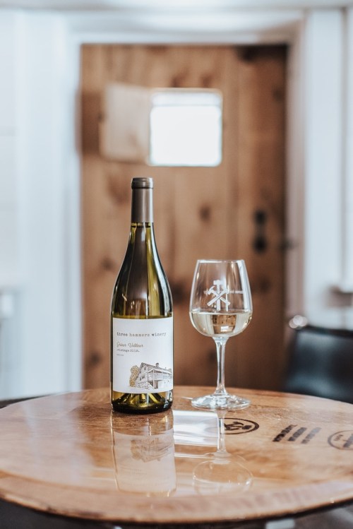 A bottle of white wine and a glass sit on a wooden table, with a rustic door in the background.