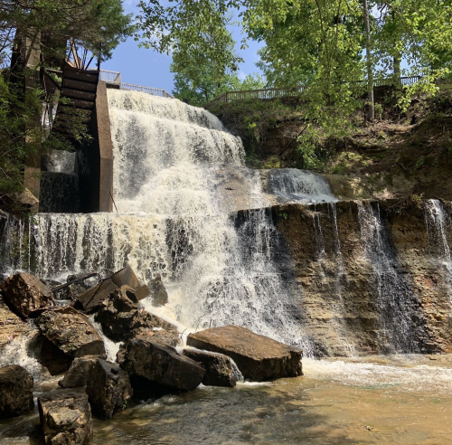 A cascading waterfall flows over rocky terrain, surrounded by lush green trees under a clear blue sky.