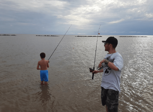 A man and a boy fishing in shallow water, with cloudy skies and a distant shoreline in the background.