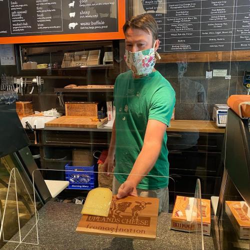 A person wearing a mask stands behind a counter, holding a wooden cheese sign in a shop.