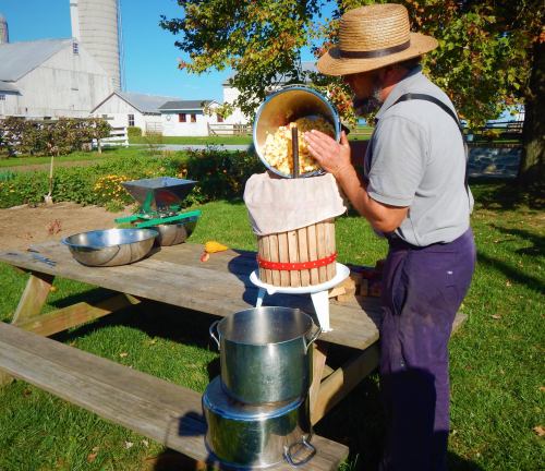 A man in a straw hat uses a wooden fruit press outdoors, surrounded by bowls and a farm setting.