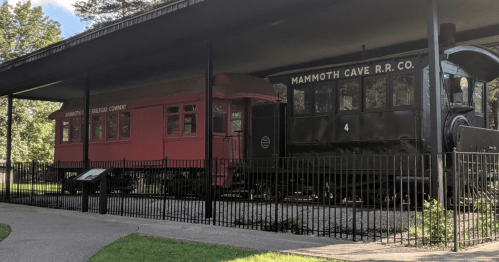 Two vintage train cars, one red and one black, displayed under a shelter with a fence around them.