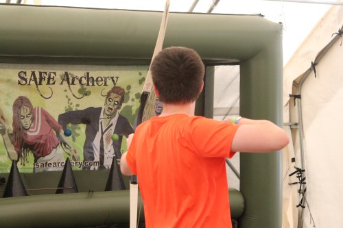 A person in an orange shirt aims an arrow at a zombie-themed archery target in a tent.