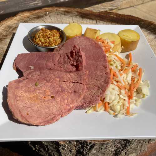 A plate with sliced corned beef, mustard, boiled potatoes, and coleslaw on a wooden surface.