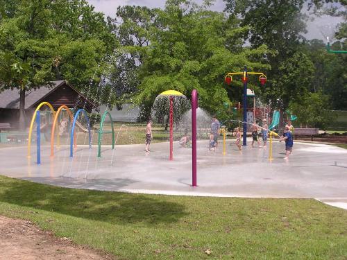 Children playing in a colorful splash pad with water features, surrounded by trees and a playground in the background.