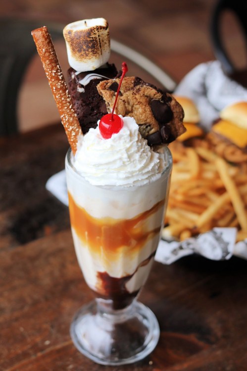 A tall sundae glass filled with ice cream, topped with whipped cream, a cherry, a cookie, a brownie, and a pretzel stick.