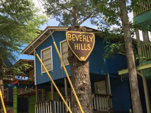 Sign reading "Beverly Hills" on a tree in front of a colorful, two-story building surrounded by greenery.
