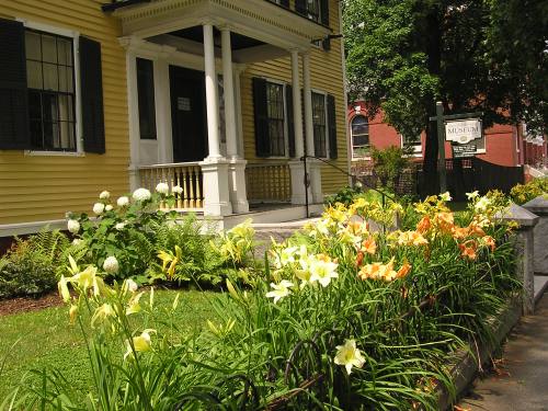 A yellow house with black shutters, surrounded by colorful flowers and greenery, with a sign for a museum nearby.