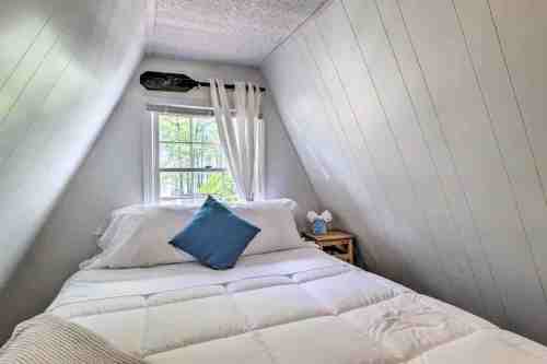 Cozy bedroom with slanted walls, a bed with white linens, a blue pillow, and a window letting in natural light.