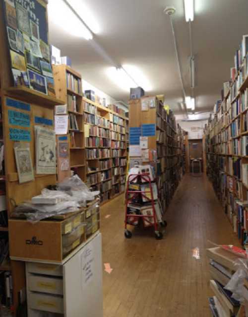 A narrow aisle lined with bookshelves in a cozy bookstore, with a cart and a chair at the far end.