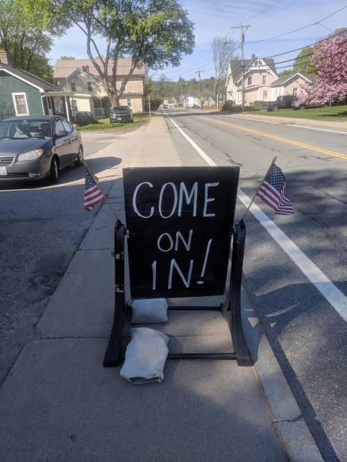 A sidewalk sign with "COME ON IN!" written in bold letters, flanked by small American flags.