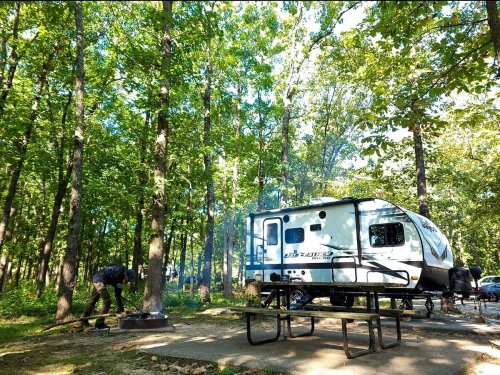 A camper parked in a wooded area, with a person tending to a fire pit and a picnic table nearby.