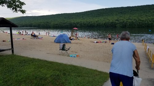 A sandy beach by a lake, with people swimming and relaxing under umbrellas, surrounded by green hills.