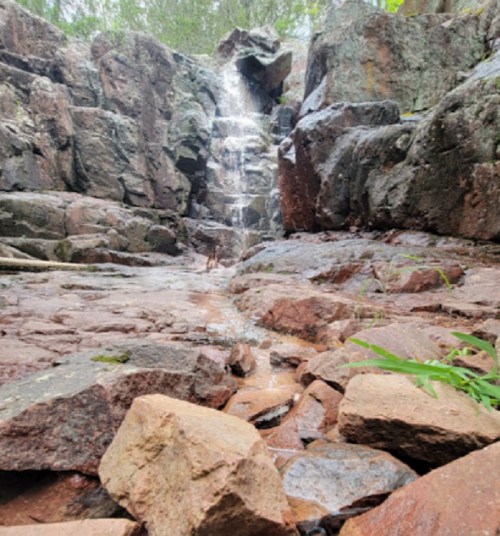 A small waterfall cascades over rocky terrain, surrounded by lush greenery and natural stone formations.