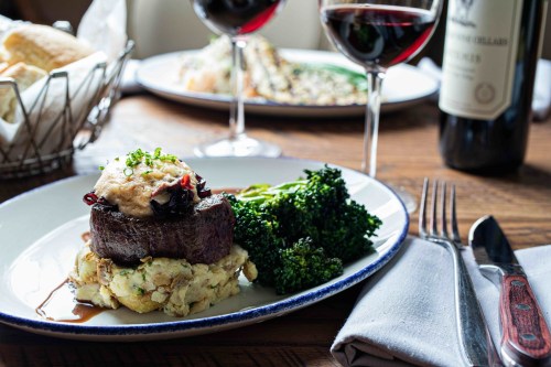 A gourmet plate featuring a filet mignon atop stuffing, accompanied by broccoli and red wine glasses in the background.