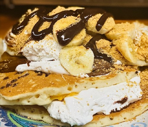 Stack of pancakes topped with whipped cream, banana slices, chocolate drizzle, and crushed graham crackers.