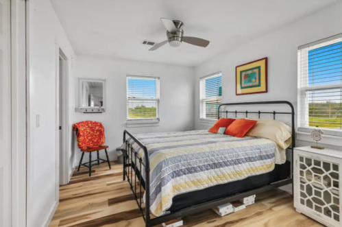 Bright bedroom with a metal bed, colorful pillows, wooden floor, and large windows letting in natural light.