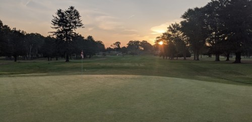 A serene golf course at sunset, with a flag on the green and trees lining the fairway.