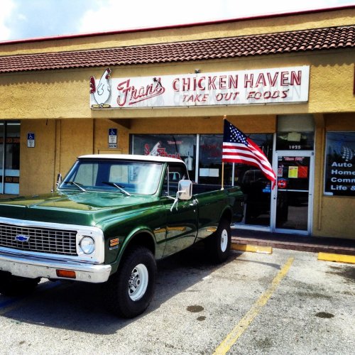 A green pickup truck parked in front of Fran's Chicken Haven, a takeout restaurant with an American flag.