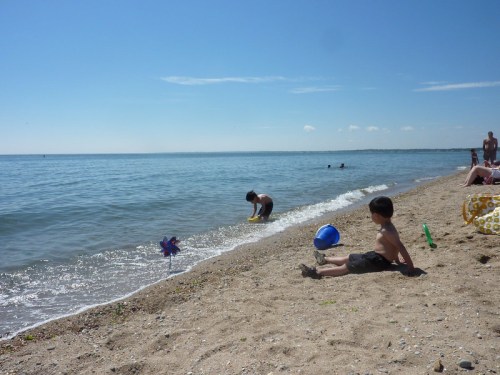 A sunny beach scene with children playing in the water and on the sand, with a clear blue sky and gentle waves.