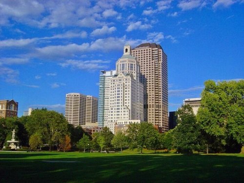 City skyline featuring modern skyscrapers against a blue sky, with green trees in the foreground.