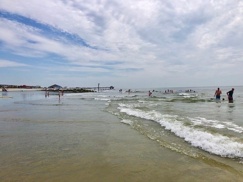 A beach scene with people swimming and walking along the shore under a cloudy sky. Waves gently lap at the sand.