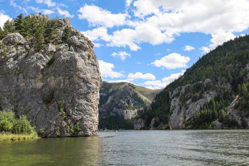 A serene river flows between towering rocky cliffs and lush green mountains under a partly cloudy sky.