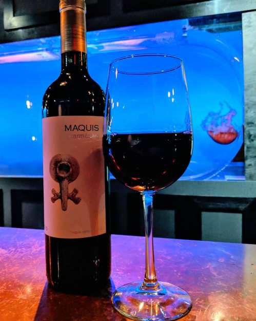 A bottle of Maquis wine next to a filled glass, with a blue-lit background featuring a fish tank.