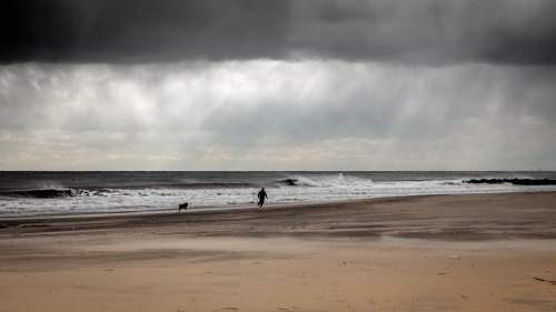 A person walks a dog along a beach under a cloudy sky with dark storm clouds and gentle waves.