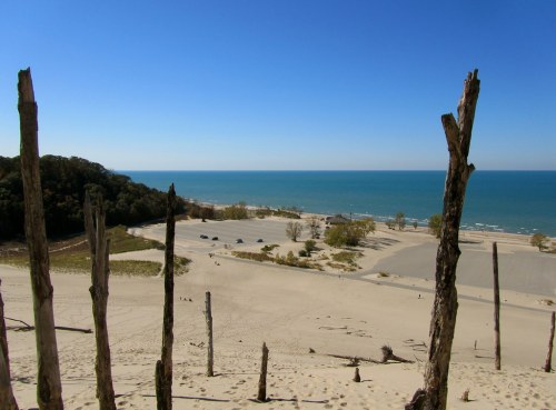 A sandy beach with a clear blue lake in the background, framed by weathered wooden posts and lush greenery.