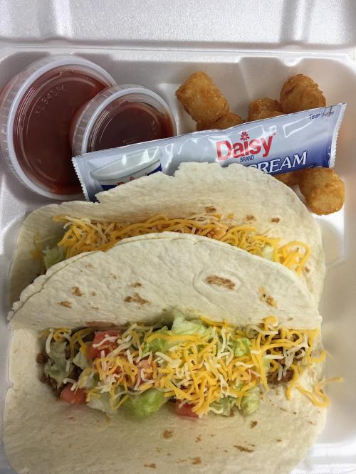 Two tacos filled with lettuce, tomatoes, and cheese, served with salsa, sour cream, and tater tots in a takeout container.