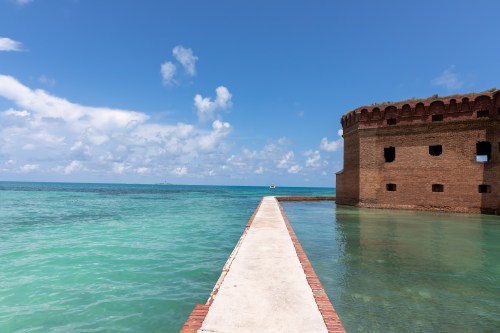 A brick fort stands by clear turquoise waters under a bright blue sky with scattered clouds. A walkway leads to the fort.